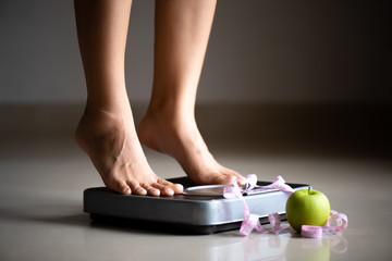 Female leg stepping on weigh scales with measuring tape and green apple. Healthy lifestyle, food...