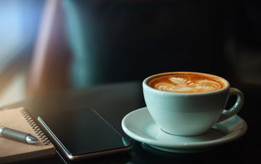 Closeup latte art coffee in white cup with smartphone, notebook and pen on black table in cafe. Vintage light
