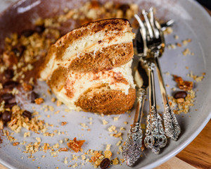 A slice of Tiramisu on a plate surrounded by coffee beans, honeycomb and nuts and some fancy cutlery