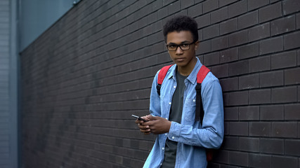 Lonely afro-american student using cellphone, bored in school backyard, outcast