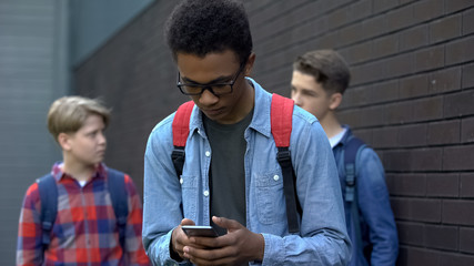 Black student reading offensive post in phone, boys mock behind, cyberbullying
