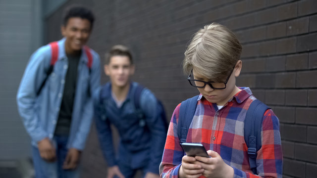 Junior student reading offensive post in phone, boys mock behind, cyberbullying