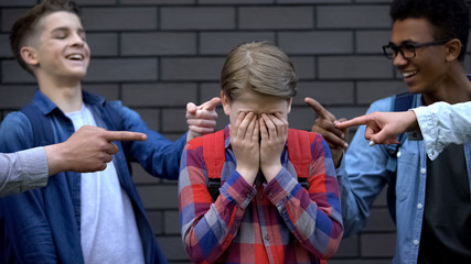 Malicious teenagers pointing fingers at boy crying, depressed victim of bullying