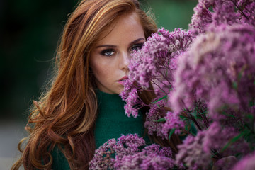 Naklejka premium Sunshine young smiling woman with red curly hair is wearing green sweater and hat in autumn park near purple bush of flowers.