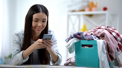 Smiling housewife chatting on smartphone near laundry basket with clothes