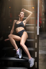 Fototapeta na wymiar Full-length portrait of a beautiful girl in black underwear looking aside while sitting on stairs. Seductive sporty brunette. Fashionable, commercial, advertising photoshoot