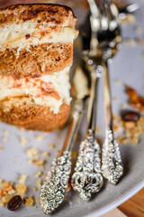 A slice of Tiramisu on a plate surrounded by coffee beans, honeycomb and nuts and some fancy cutlery
