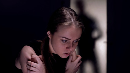 Scared crying woman sitting alone in room, cruel tyrant husband shadow outside