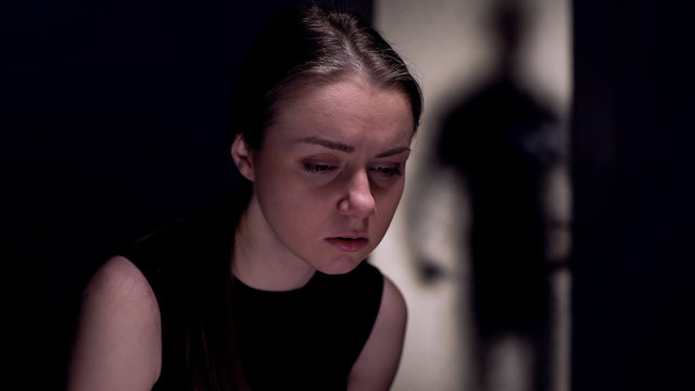 Sad woman looking down, dangerous man shadow at door, human trafficking victim