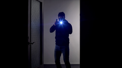 Masked man with gun and flashlight standing at door, police service, crime scene
