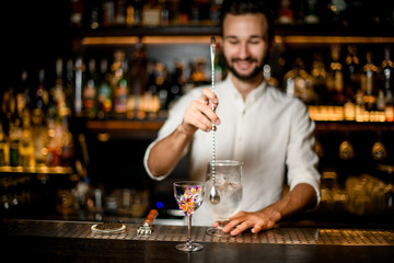 Smiling bartender stirring a cocktail with a spoon in the focus foreground of flower decorated glass