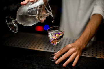 Professional bartender pouring a cocktail from the measuring cup with a strainer to a glass decorated with flowers