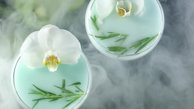 glass glasses with a blue cocktail, a white Orchid flower and a sprig of tarragon stand on a black stone stand. festive drinks are shrouded in smoke. Top view. shooting with Studio lighting