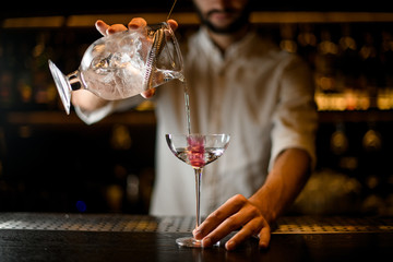 Professional male bartender pouring a cocktail from the measuring cup with a strainer to a glass with pink ice cube