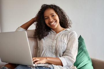 People, technology and communication. Pretty girl with voluminous Afro hairstyle sitting on couch...