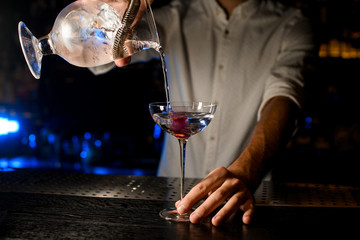 Bartender pouring a cocktail from the measuring cup with a strainer to a glass with pink ice cube