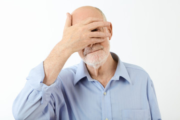 Portrait of elderly retired Caucasian man in blue shirt holding hand on his face, covering eyes and...