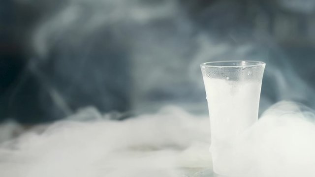 a frosted glass of vodka sits empty on a frozen glass tray. steam escapes from it and spreads across the wooden table. Panorama shooting.