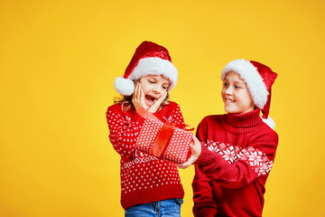 Happy boy in Santa hat giving Christmas gift box to excited girl in red sweater with deer on yellow background