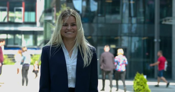Attractive young blonde woman wearing smart business attire, stands in front of office buildings, smiling as the breeze blows her hair around