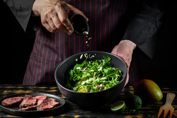 Cooking fresh diet keto salad with beef meat, arugula, fruits and nuts by chef hands on black table background.