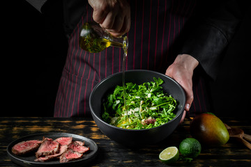 Cooking fresh diet keto salad with beef meat, arugula, fruits and nuts by chef hands on black table background.