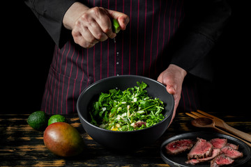Cooking fresh diet keto salad with beef meat, arugula, fruits and nuts by chef hands on black table background.