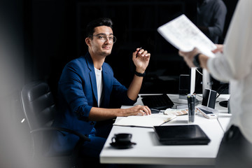 The architect in glasses dressed in a blue jacket sits at the desk in front of the computer and looks and the other man