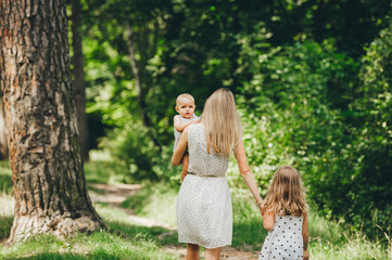 Beautiful long-haired mother in a summer dress in sunny forest walks with his beautiful daughters. Back view.