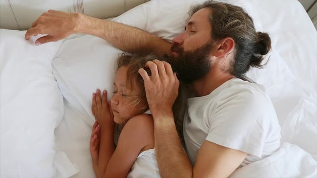Little daughter sleeping with her bearded father while he is stroking her head. Father and cute daughter lying together on the bed in bedroom at home