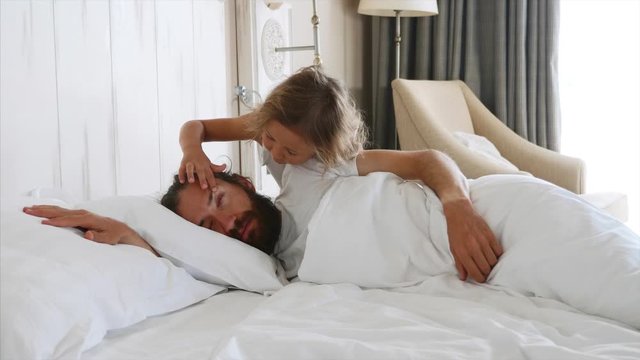 Little cute daughter comes to sleeping father at morning, and opens his eyes by fingers for wakes him. Dad covering his head by pillow. Family at home concept.
