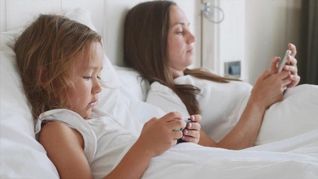 Young mother with little cute daughter are using gadgets lying on a white bed at domestic bedroom. Concept modern family during weekend at home with mobile phones in hands, side view..