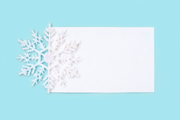 Fototapeta na wymiar White paper card decorated with snowflakes on light blue background. New Year, Christmas and winter concept. Flat lay, top view, free copy space.