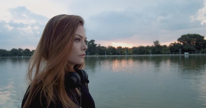 Beautiful young woman with wireless headphones standing on a river shore at sunset
