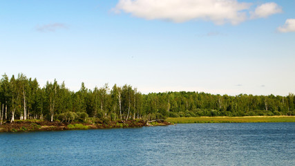 Lake and wood on the shore against blue sky and white clouds on a summer day