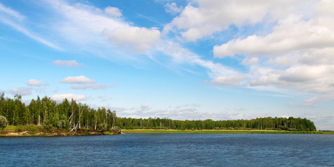 Lake and wood on the shore against blue sky and white clouds on a summer day