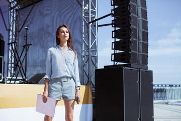 Installation of stage equipment and preparing for a live concert open air. Event manager portrait. Summer music city festival. Young serious woman stand and work with her laptop near the stage... - 287022395