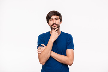 Thoughtful handsome brunet man in a blue shirt holding arms folded over his chest and keeping one hand close to face standing isolated over white background.