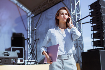 Installation of stage equipment and preparing for a live concert open air. Event manager portrait. Summer music city festival. Young serious woman stand and work with her laptop near the stage...