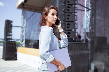 Installation of stage equipment and preparing for a live concert open air. Event manager portrait. Summer music city festival. Young serious woman stand and work with her laptop near the stage... - 287022744