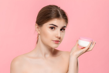 Beautiful girl holding tube of cream to moisturize skin. Model looks at the camera isolated over pink background. Concept of beauty and health treatment.