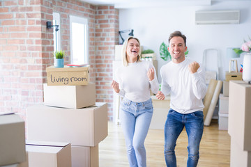 Young beautiful couple standing at new home around cardboard boxes very happy and excited doing winner gesture with arms raised, smiling and screaming for success. Celebration concept.