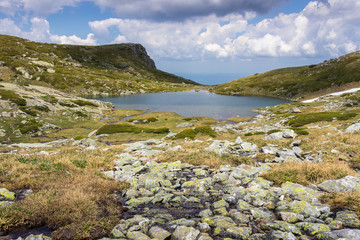Naklejka na ściany i meble Foreground rocks covered by yellow moss, blue lake, rocky peak and amazing sunlit landscape of Rila mountain in Bulgaria