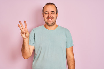 Fototapeta premium Young man wearing blue casual t-shirt standing over isolated pink background showing and pointing up with fingers number three while smiling confident and happy.