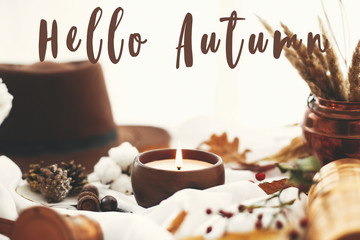 Hello Autumn text, fall greeting sign on candle, berries, fall leaves, herbs, acorns, nuts and brown hat on white fabric. Hygge lifestyle, cozy inspiration
