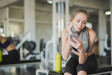 Young woman take a rest after workout at gym which looking and playing a smart phone. Woman sitting and searching in smart phone at the fitness room near the big mirror.