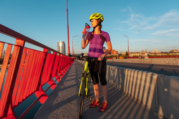 Sporty girl standing in the sunset light with her bike and drinking water