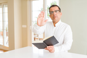 Middle age man reading a book at home wearing glasses with open hand doing stop sign with serious and confident expression, defense gesture