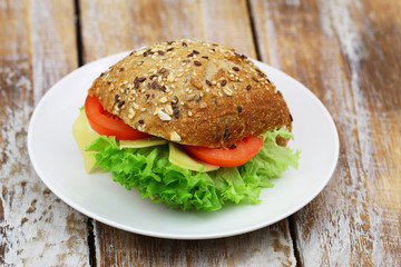 Whole grain roll with cheese, lettuce and tomato on white plate on rustic wooden surface