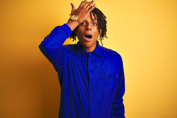 Afro worker man with dreadlocks wearing mechanic uniform over isolated yellow background surprised with hand on head for mistake, remember error. Forgot, bad memory concept.